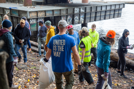 A beach full of people cleaning. A man in a blue shirt that says United is the Way stands central in the image