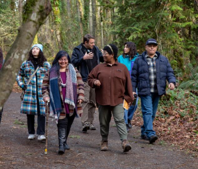 A group of people walk through a forest while two women in front smile and talk