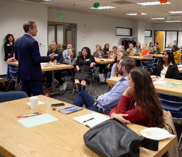 A group of people sit listening to a speaker in a plain office room